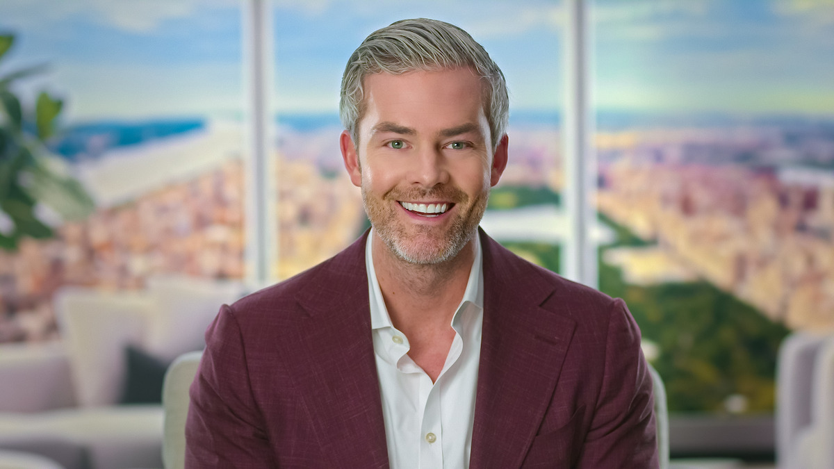 Smiling man with gray hair in a maroon suit sits indoors with a bright, modern cityscape visible through large windows behind him; the setting is professional and upbeat.