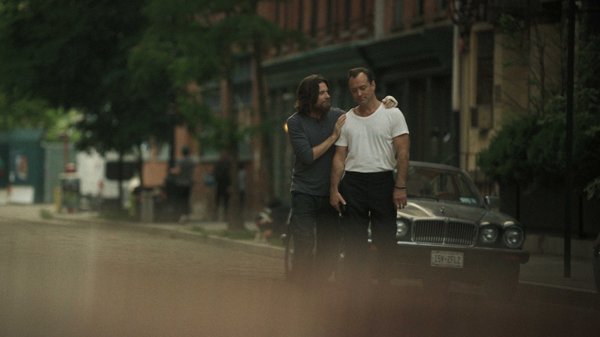 Two men walk closely together on a quiet city street beside a vintage car, surrounded by brick buildings and greenery on a cloudy day.