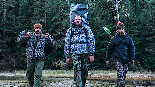 Three 'Outlast' Season 2 contestants walk towards the camera with their equipment and supplies.