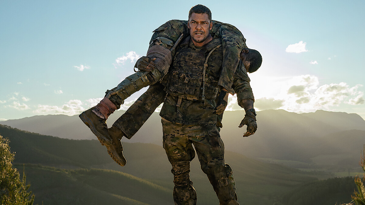 Soldier in camouflage uniform carrying another soldier over his shoulders, standing outdoors in a mountainous landscape with a bright sky in the background.
