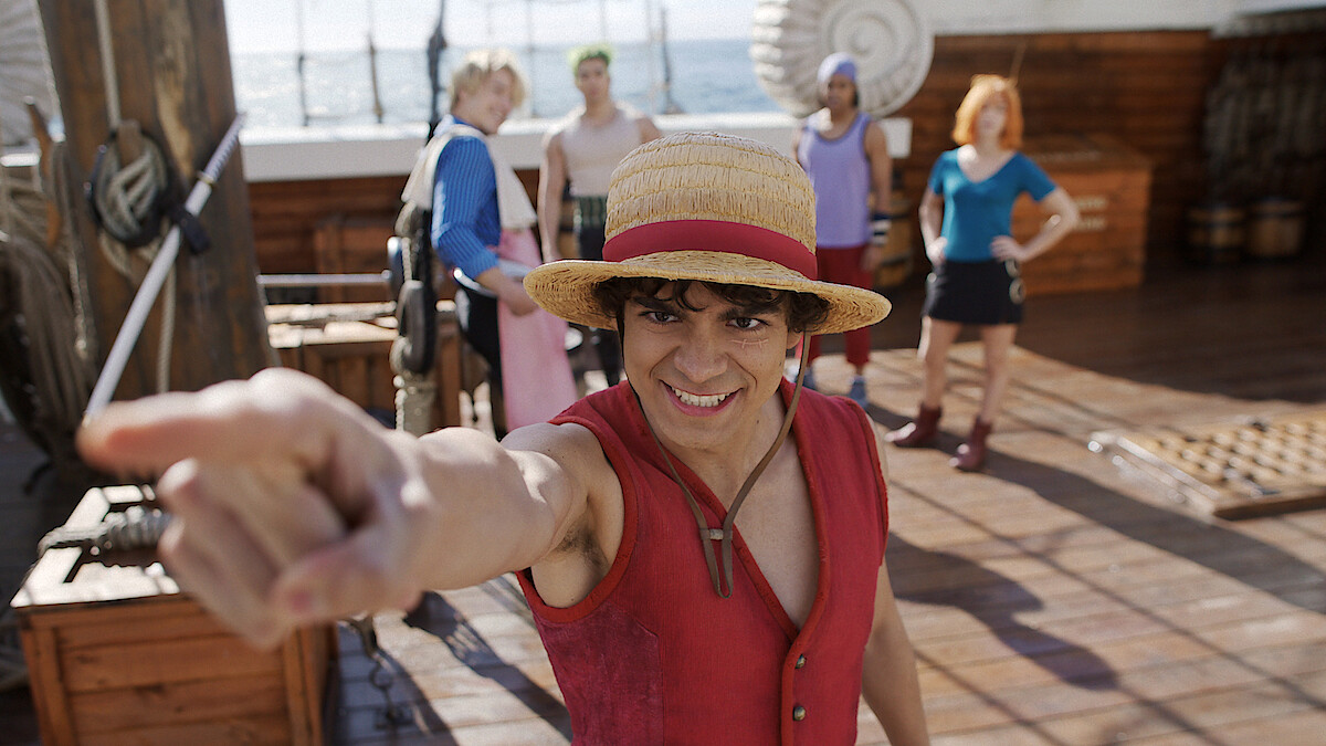 Person in a straw hat and red vest pointing forward on a wooden ship deck, with ocean in the background and four people standing behind, dressed in colorful outfits, under bright daylight.
