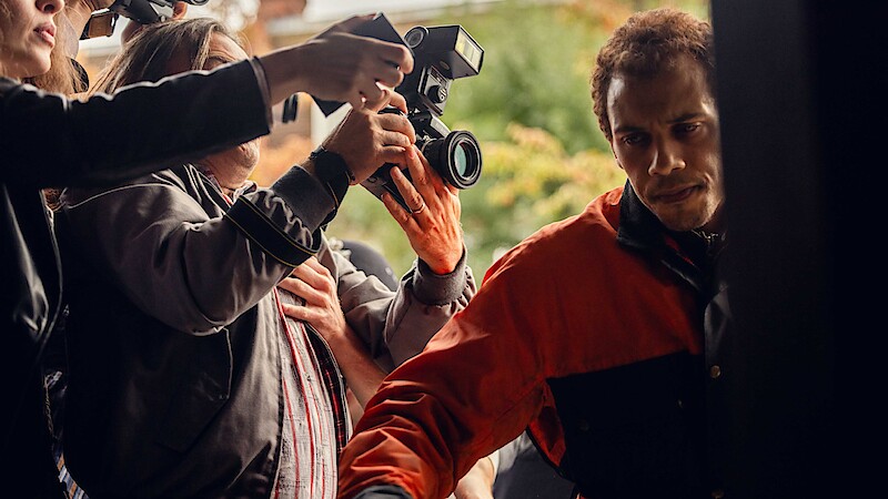 A man in a red and black jacket tries to move through a crowd outside as people with cameras and microphones surround him, suggesting a media or paparazzi situation.