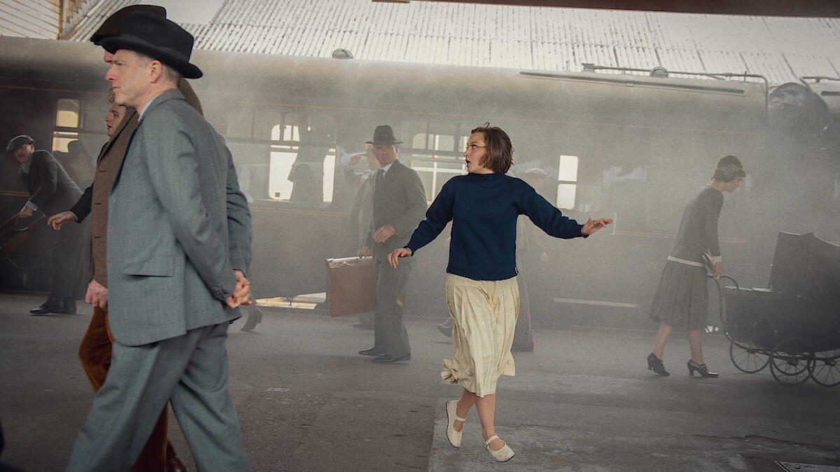 A woman in a skirt and sweater looks around anxiously at a vintage train station as people in old-fashioned clothing walk by, with steam and luggage visible in the background.