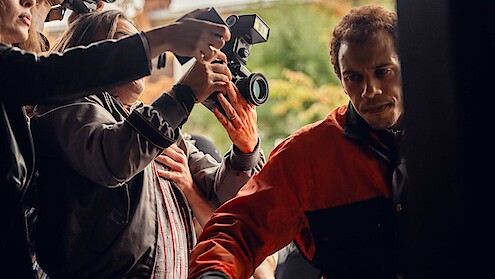 A man in a red and black jacket tries to move through a crowd outside as people with cameras and microphones surround him, suggesting a media or paparazzi situation.