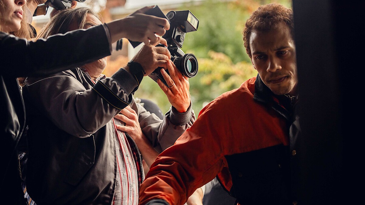 A man in a red and black jacket tries to move through a crowd outside as people with cameras and microphones surround him, suggesting a media or paparazzi situation.