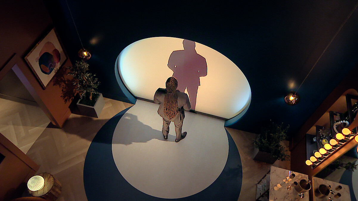 Dr Vic St John standing alone in a modern, dimly lit room with blue walls, casting a large shadow onto a circular white light on the wall. Decorative plants, artwork, and a set dining table create a stylish, intimate environment.