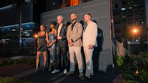 Five well-dressed people posing together on an outdoor evening event stage, city buildings and greenery in the background, creating a formal and celebratory mood.