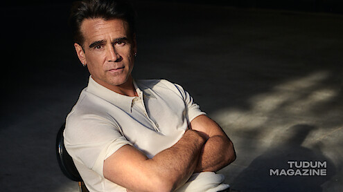 Colin Farrell leans back in a rolling chair in a white polo and black pants. 