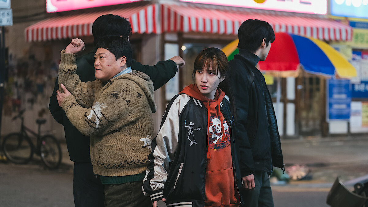 Four people stand on a city street at night near shops with striped awnings; one person looks concerned, two face away, and the street is dimly lit with visible store signs and urban elements.